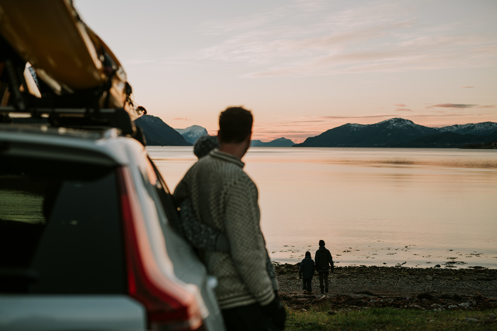 Mann og dame lener seg mot bilen, mens de ser på barna som leker nede på standen ved solnedgang.